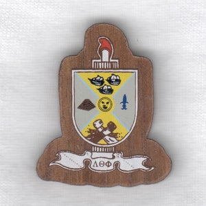 May include: Wooden plaque with a shield design. The shield features a torch, ships, a pyramid, and other symbols. A banner at the bottom displays Greek letters. The plaque has a brown border and is set against a white background.