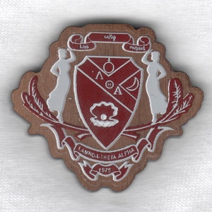 May include: A wooden Lambda Theta Alpha crest with a red shield, white accents, and the year 1975. The crest features the words "Love, Unity, Respect" and stylized figures.