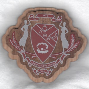 May include: A wooden crest with a red shield featuring the letters "LΘA" and a pearl. The shield is surrounded by two figures and a banner that reads "Lambda-Theta-Alpha 1975". The crest is decorated with a wooden grain pattern.
