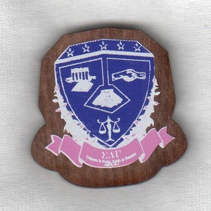 May include: A wooden shield-shaped pin with a blue and white crest featuring a building, a hand, and a scale. The shield is surrounded by a white wreath and a pink ribbon with the text "ΣΑΓ Culture & Pride in Excellence" and the letters "ΣΑΓ".