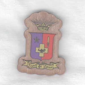 May include: A wooden emblem with a shield design featuring a star, a sword, and a lamp with a cross. The shield is divided into purple and red sections. A gold crown sits above the shield. The emblem has Greek text on a banner.