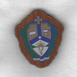 May include: A wooden shield-shaped plaque with a crest featuring a white lamb, a gold lamp, a blue star, and a sunburst. The crest is surrounded by a green wreath and the words "Lux et Veritas" are inscribed at the bottom.