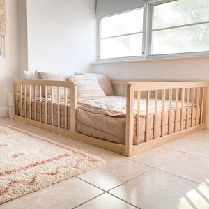 Op de afbeelding: Een lichtgekleurd houten peuterbed met een lattenontwerp. Het bed is opgemaakt met een blush-kleurige quilt en kussens. Een cr&egrave;mekleurig tapijt met een roze ontwerp ligt voor het bed. Een geweven wandkleed en een mand zijn ook in de kamer.