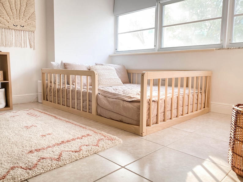 Op de afbeelding: Een lichtgekleurd houten peuterbed met een lattenontwerp. Het bed is opgemaakt met een blush-kleurige quilt en kussens. Een cr&egrave;mekleurig tapijt met een roze ontwerp ligt voor het bed. Een geweven wandkleed en een mand zijn ook in de kamer.