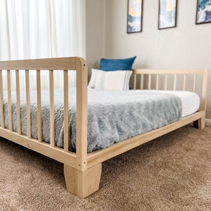 Op de afbeelding: Een lichtgekleurd houten bedframe met een lattenbodem voor hoofd- en voeteneinde. Het bed heeft een lichtblauwe pluche deken en witte lakens. Een blauw kussen en een gestreept kussen liggen op het bed. Het bed staat op een bruin tapijt.