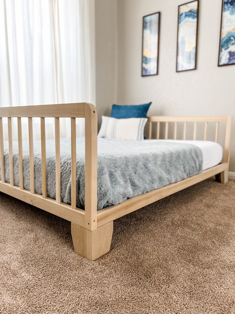 Op de afbeelding: Een lichtgekleurd houten bedframe met een lattenbodem voor hoofd- en voeteneinde. Het bed heeft een lichtblauwe pluche deken en witte lakens. Een blauw kussen en een gestreept kussen liggen op het bed. Het bed staat op een bruin tapijt.