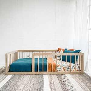 K&ouml;nnte beinhalten: Ein Holzkinderbett mit einer t&uuml;rkisfarbenen Decke und Kissen. Die Bettw&auml;sche umfasst eine orangefarbene Decke und ein gemustertes Laken mit F&uuml;chsen und floralen Designs. Das Bett steht vor einer wei&szlig;en Backsteinmauer und einem gemusterten Teppich.