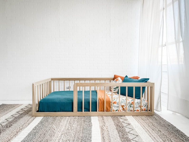 K&ouml;nnte beinhalten: Ein Holzkinderbett mit einer t&uuml;rkisfarbenen Decke und Kissen. Die Bettw&auml;sche umfasst eine orangefarbene Decke und ein gemustertes Laken mit F&uuml;chsen und floralen Designs. Das Bett steht vor einer wei&szlig;en Backsteinmauer und einem gemusterten Teppich.