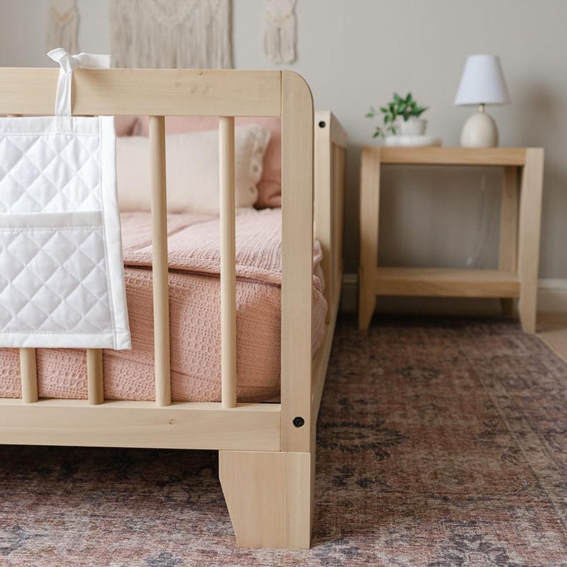 May include: A light wood toddler bed with a white quilted storage bag hanging on the side. The bed has a pink blanket and a white pillow. The bed is on a patterned rug.