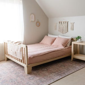 May include: A light wood toddler bed with a pink quilted bedspread and two pink pillows. The bed has a side rail with pockets for storage. The bed is in a bedroom with a light gray wall and a rug with a pink and gray pattern.