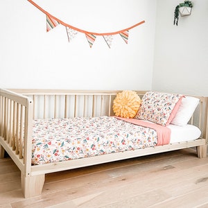 K&ouml;nnte beinhalten: Ein helles Holzkinderbett mit einem Bettw&auml;sche-Set mit Blumenmuster. Die Bettw&auml;sche hat einen wei&szlig;en Hintergrund mit roten, gelben und gr&uuml;nen Blumenmustern. Ein gelbes bl&uuml;tenf&ouml;rmiges Kissen und eine rosa Decke befinden sich ebenfalls auf dem Bett.
