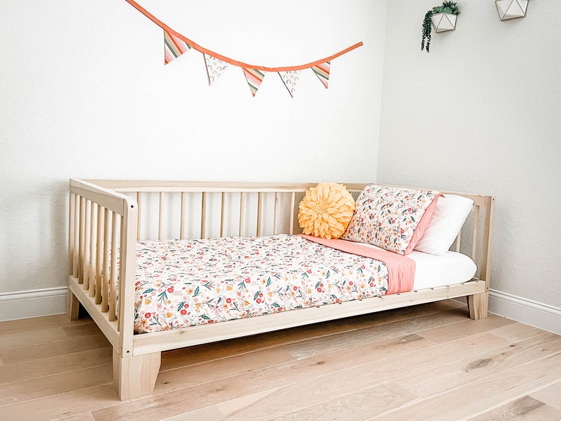 K&ouml;nnte beinhalten: Ein helles Holzkinderbett mit einem Bettw&auml;sche-Set mit Blumenmuster. Die Bettw&auml;sche hat einen wei&szlig;en Hintergrund mit roten, gelben und gr&uuml;nen Blumenmustern. Ein gelbes bl&uuml;tenf&ouml;rmiges Kissen und eine rosa Decke befinden sich ebenfalls auf dem Bett.