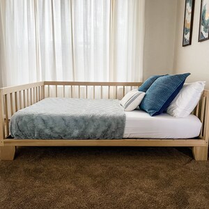 May include: A wooden daybed with a light blue blanket and blue and white pillows. The daybed is in a room with white curtains and beige walls.
