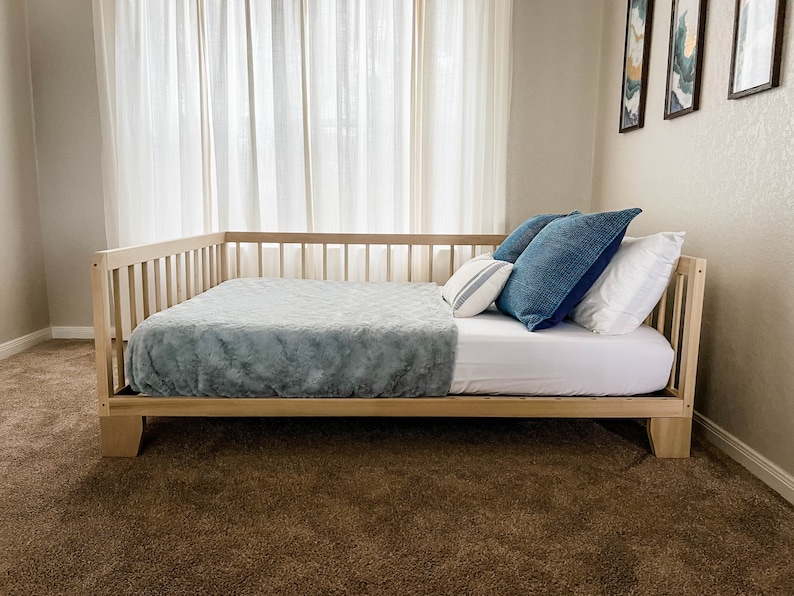 May include: A wooden daybed with a light blue blanket and blue and white pillows. The daybed is in a room with white curtains and beige walls.