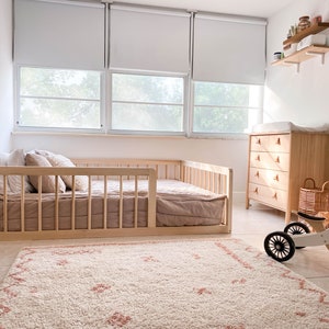 Op de afbeelding: Een lichte kinderkamer met een licht houten bed met spijlen. Er is een commode van licht hout, een tapijt met wit-roze patroon en een houten driewieler met mand. De kamer is licht en luchtig.