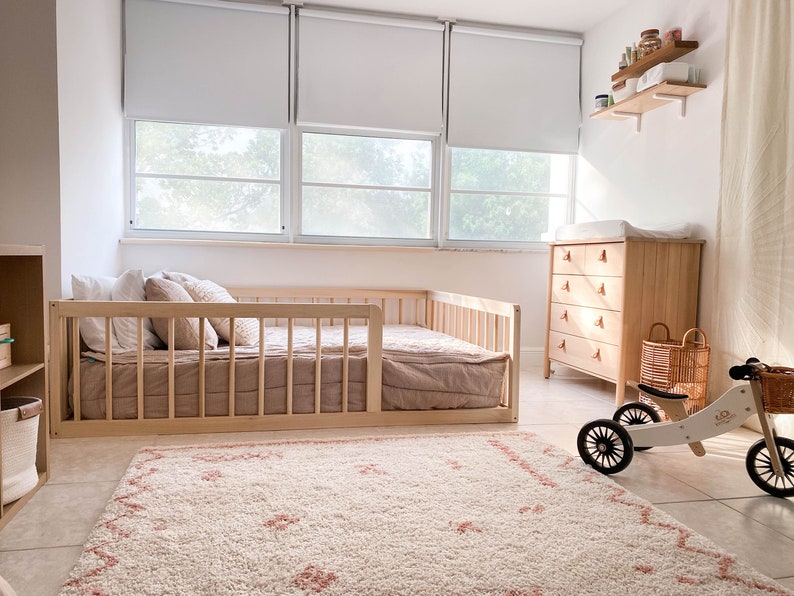 Op de afbeelding: Een lichte kinderkamer met een licht houten bed met spijlen. Er is een commode van licht hout, een tapijt met wit-roze patroon en een houten driewieler met mand. De kamer is licht en luchtig.