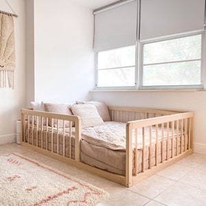 Op de afbeelding: Een peuterbed met een licht houten frame, gevuld met beige en roze beddengoed. Het bed staat in een lichte kamer met een vloerkleed en wanddecoratie. Afmetingen van het bed: ongeveer 140 cm x 70 cm.