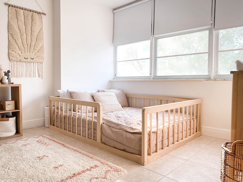 Op de afbeelding: Een peuterbed met een licht houten frame, gevuld met beige en roze beddengoed. Het bed staat in een lichte kamer met een vloerkleed en wanddecoratie. Afmetingen van het bed: ongeveer 140 cm x 70 cm.