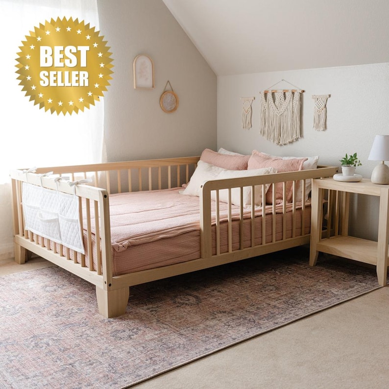 May include: A light wood toddler bed with a pink bedspread and white guard rails. The bed is on a pink and gray patterned rug. There is a small wooden nightstand next to the bed.