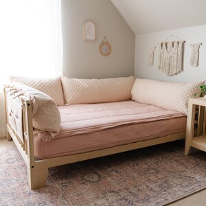 May include: A light wood daybed with a pink quilted mattress and matching pillows. The daybed is in a room with a light gray wall and a rug with a brown and pink pattern.