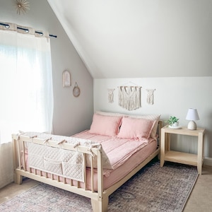 K&ouml;nnte beinhalten: Ein helles Holzbett mit rosa Bettw&auml;sche und Kissen. Ein gestepptes Kopfteil mit Taschen ist befestigt. Das Bett steht auf einem gemusterten Teppich. Ein kleiner Nachttisch mit Lampe und Makramee-Wandbeh&auml;ngen ist ebenfalls zu sehen.