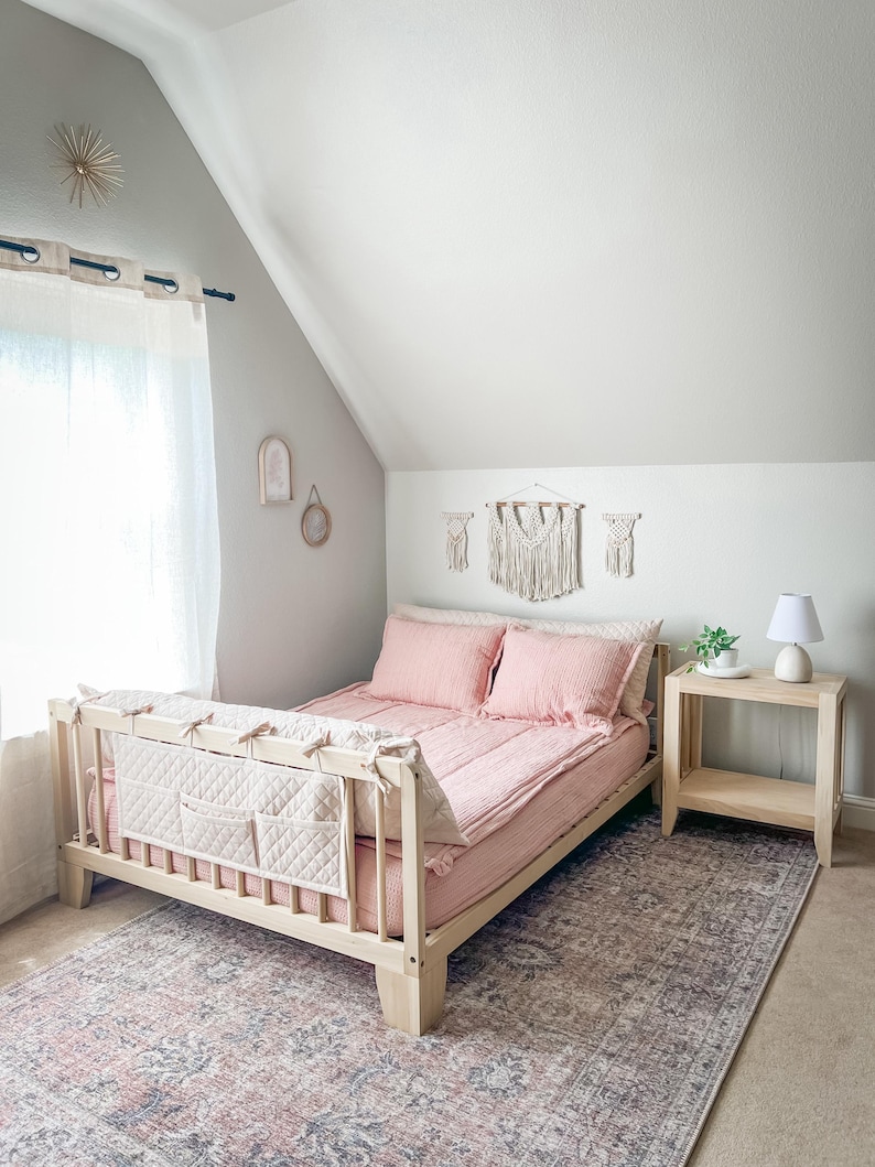 K&ouml;nnte beinhalten: Ein helles Holzbett mit rosa Bettw&auml;sche und Kissen. Ein gestepptes Kopfteil mit Taschen ist befestigt. Das Bett steht auf einem gemusterten Teppich. Ein kleiner Nachttisch mit Lampe und Makramee-Wandbeh&auml;ngen ist ebenfalls zu sehen.