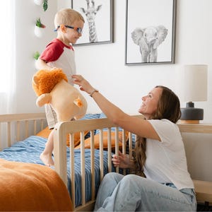May include: A young boy wearing glasses and a red and white shirt is standing on a wooden toddler bed. He is holding a stuffed lion. A woman is sitting on the bed, reaching out to the boy. The bed has a blue and white striped sheet and a brown blanket.
