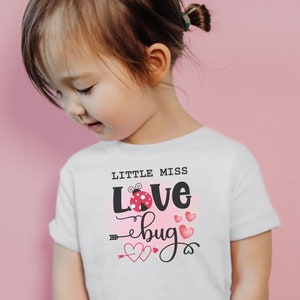 Pode incluir: Uma camiseta branca com um gráfico de joaninha rosa e preta e o texto "Little Miss Love Bug" com corações rosas.