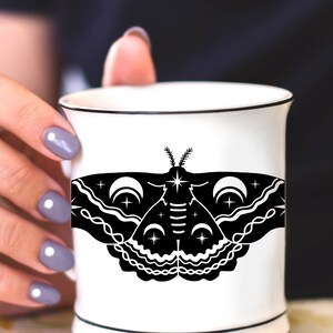 Puede incluir: Una taza de cer&aacute;mica blanca con una ilustraci&oacute;n en blanco y negro de una polilla con lunas crecientes y estrellas. La polilla tiene un dise&ntilde;o detallado con l&iacute;neas y patrones intrincados.