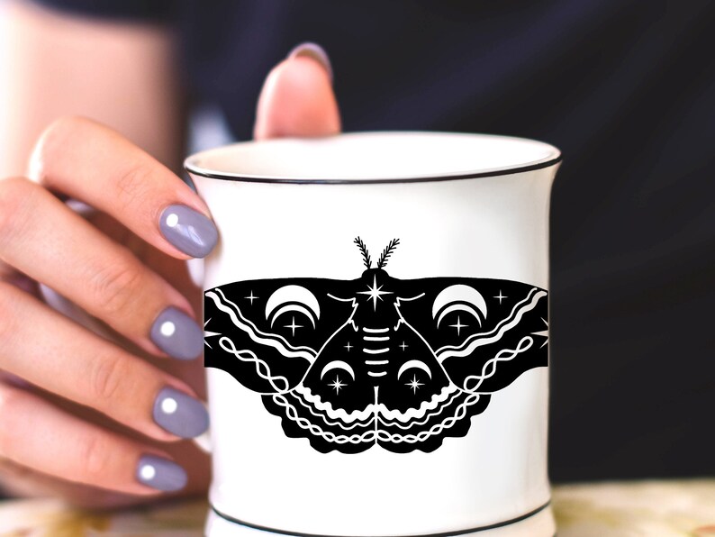 Puede incluir: Una taza de cer&aacute;mica blanca con una ilustraci&oacute;n en blanco y negro de una polilla con lunas crecientes y estrellas. La polilla tiene un dise&ntilde;o detallado con l&iacute;neas y patrones intrincados.