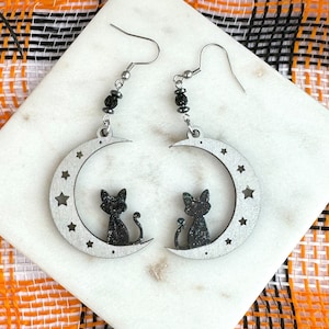 May include: A pair of silver crescent moon earrings with black glitter and two black cats sitting on the moon. The earrings are on a white marble surface.