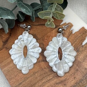 May include: A pair of white, textured, oval-shaped earrings with a silver post. The earrings have a scalloped edge design and a central oval opening. They are displayed on a wooden surface.