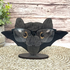 May include: A black wooden bat with a pair of glasses perched on its face. The bat has a black and white painted design and is standing on a wooden base.