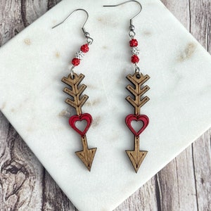 May include: A pair of wooden arrow earrings with red hearts and white and red beads. The earrings are on a white marble surface.