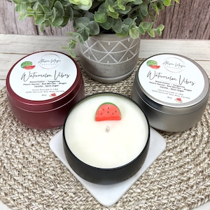May include: Three 8 oz soy wax candles in red, silver, and black tins with lids. Each candle has a watermelon slice on top and the label "Watermelon Vibes" with a list of scents: Watermelon, Tangerine, Peach Nectar, Red Berries, Muget, Vanilla, Spun Sugar. Hand poured in the USA.