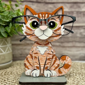 May include: A wooden cat-shaped glasses holder in orange and white stripes, with green eyes and a pink nose. The cat is wearing black-framed glasses. The holder sits on a gray rectangular base, perfect for a desk or nightstand.