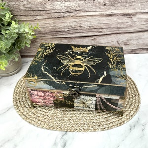 May include: A black wooden box with a gold bee design and a crown on top. The box is decorated with pink and gold floral patterns and has a hinged lid. The box is sitting on a woven straw mat.