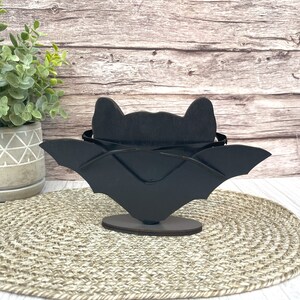 May include: Black wooden bat silhouette with wings outstretched, standing on a round base. The bat is a decorative object for Halloween.