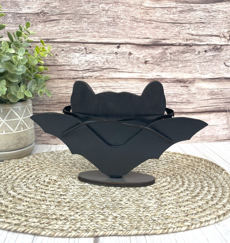 May include: Black wooden bat silhouette with wings outstretched, standing on a round base. The bat is a decorative object for Halloween.
