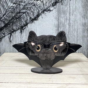 May include: A black bat-shaped glasses holder with gold-rimmed glasses resting on it. The holder has a black base and is made of wood.