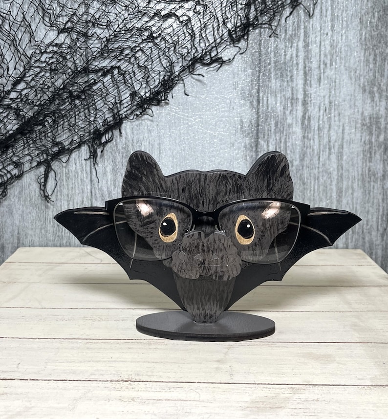 May include: A black bat-shaped glasses holder with gold-rimmed glasses resting on it. The holder has a black base and is made of wood.