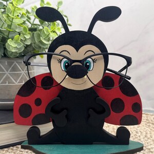 May include: A wooden ladybug-shaped eyeglasses holder. The black ladybug has red wings with black spots, blue eyes, and is wearing black-framed glasses. The holder is on a green base and is designed to hold eyeglasses.