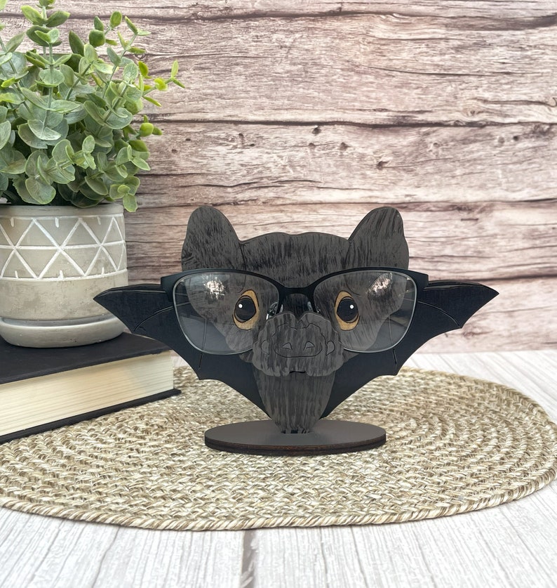 May include: A black wooden bat with glasses perched on its nose. The bat is wearing a pair of black framed glasses. The bat is standing on a wooden base. The bat is a cute and whimsical way to hold your glasses.