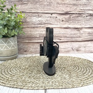 May include: A wooden glasses holder with a black finish. The holder has a rectangular base and a curved top that holds the glasses. The holder is sitting on a woven mat.