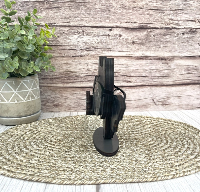 May include: A wooden glasses holder with a black finish. The holder has a rectangular base and a curved top that holds the glasses. The holder is sitting on a woven mat.