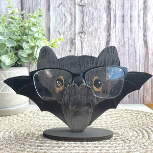May include: A black wooden bat-shaped eyeglasses holder with a pair of black glasses on it. The holder has a textured wood grain finish and a small base. The holder is on a brown woven surface.
