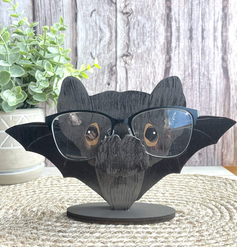 May include: A black wooden bat-shaped eyeglasses holder with a pair of black glasses on it. The holder has a textured wood grain finish and a small base. The holder is on a brown woven surface.