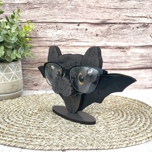 May include: A black wooden bat with glasses perched on its nose. The bat has a small, rounded body and large, pointed wings. The glasses are black with a dark gray frame.