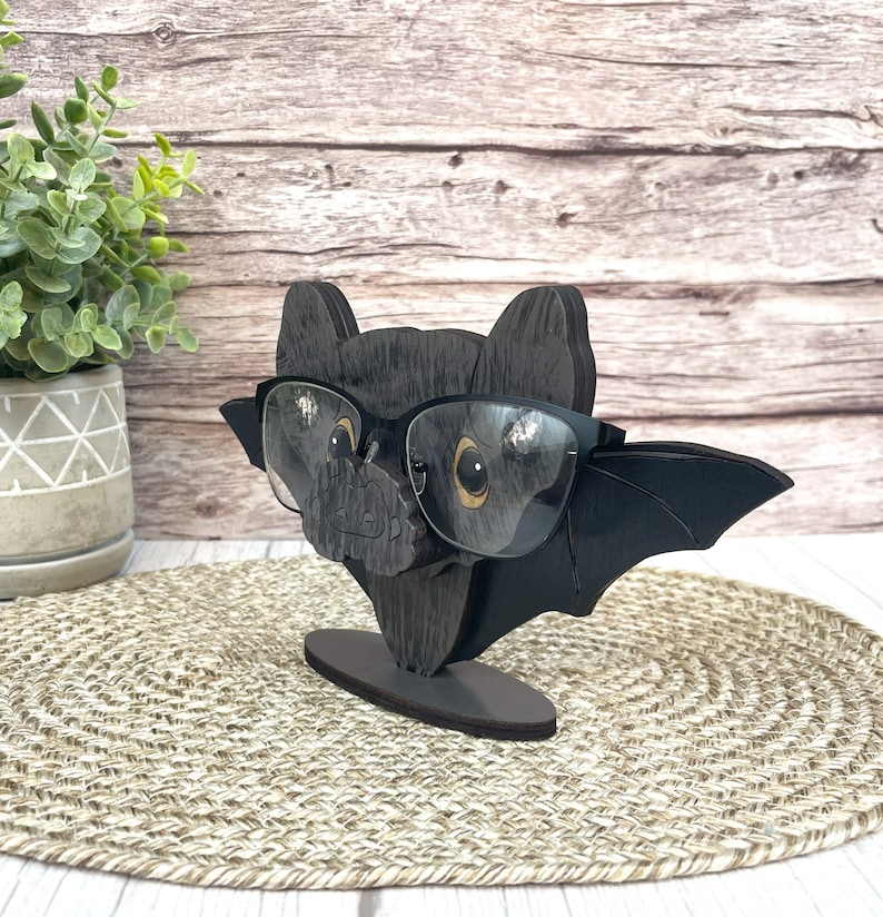 May include: A black wooden bat with glasses perched on its nose. The bat has a small, rounded body and large, pointed wings. The glasses are black with a dark gray frame.
