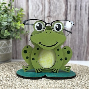 May include: A green frog figurine with black eyes and a wide smile, wearing black glasses, sits on a green lily pad. The frog is holding a pair of glasses in its hands.